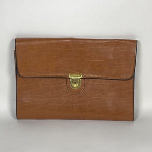 May include: A rectangular, brown leather briefcase with a flap closure and a gold-colored clasp. The briefcase has a textured surface and black trim. The design is simple and classic, suitable for carrying documents or a laptop.