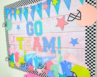 Puede incluir: Un tablero de anuncios rosa con un borde a cuadros y un fondo de tablones de madera. El tablero de anuncios está decorado con estrellas de colores, banderines y recortes con temática de fútbol. El texto "GO TEAM!" se muestra en letras grandes y coloridas. También hay banderines más pequeños con las palabras "Super", "Champions", "Fight" y "Hooray".