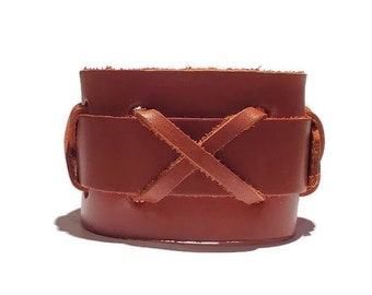 May include: A wide, brown leather cuff bracelet. The bracelet features a wrap-around design with an X-shaped leather tie detail. The leather has a natural, textured appearance, and the bracelet is set against a white background.