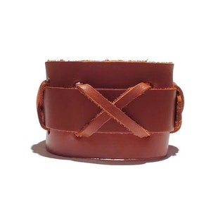 May include: A wide, brown leather cuff bracelet. The bracelet features a wrap-around design with an X-shaped leather tie detail. The leather has a natural, textured appearance, and the bracelet is set against a white background.