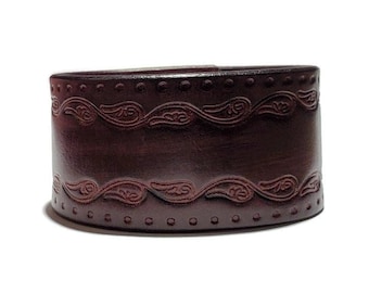 May include: A dark brown leather cuff bracelet with a decorative embossed pattern. The pattern features a repeating design of curved lines and swirls.