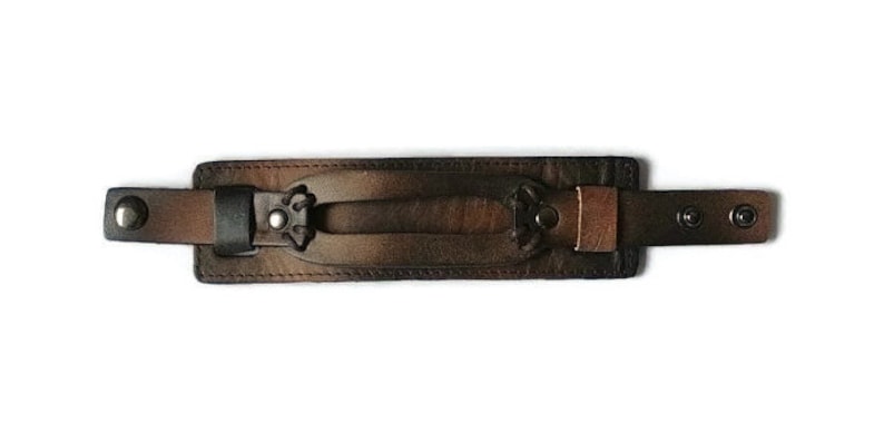 May include: A brown leather wrist cuff with a central handle and decorative metal accents. The cuff features adjustable straps with snap closures and is designed for a secure fit. The leather has a distressed finish.