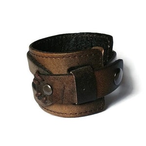 May include: A brown leather cuff bracelet with a dark brown strap and a metal snap closure. The cuff has a distressed finish and visible stitching. The bracelet is designed to wrap around the wrist.