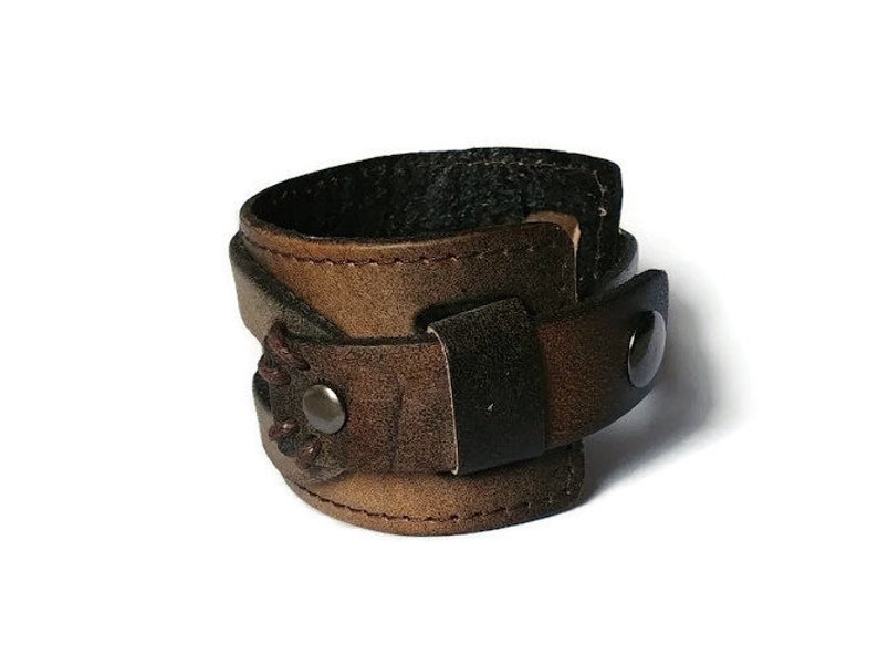 May include: A brown leather cuff bracelet with a dark brown strap and a metal snap closure. The cuff has a distressed finish and visible stitching. The bracelet is designed to wrap around the wrist.
