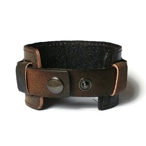 May include: A brown leather cuff bracelet with a distressed finish. The bracelet features a wide band with a black interior and a buckle closure with two metal snap buttons. The leather has a gradient of brown tones.