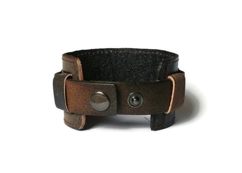 May include: A brown leather cuff bracelet with a distressed finish. The bracelet features a wide band with a black interior and a buckle closure with two metal snap buttons. The leather has a gradient of brown tones.