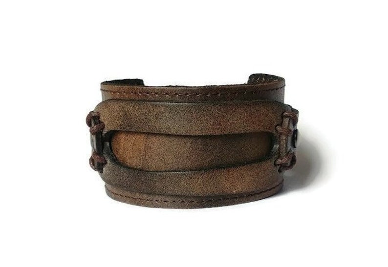 May include: A brown leather cuff bracelet with a distressed finish. The wide bracelet features three stacked bands and decorative stitching on the sides. The cuff is designed to be worn around the wrist.