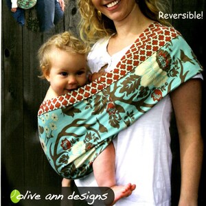 May include: A turquoise and brown patterned baby sling with a red trim. The sling is being worn by a woman with blonde hair. The sling is reversible and has the text "Sling Buddies OAD 62 Safe & Secure Baby Slings for the Whole Family Reversible!" printed on it.