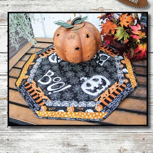 Könnte beinhalten: Eine schwarz-weiße Tischunterlage mit einem Spinnennetz-Design und dem Wort "Boo!" in weißer Schrift. Die Unterlage ist von einem schwarz-orangen gestreiften Rand umgeben. Die Unterlage hat einen Durchmesser von 56 cm und ist achteckig. Die Unterlage liegt auf einem Holztisch mit einem Metallkürbis und Herbstblättern.