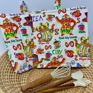 Puede incluir: Un salvamanteles acolchado blanco con diseños coloridos de teteras y tazas, con las palabras "Twist & Spout", "Read My Leaves", "Royal Tea" y "Cups". Un batidor, una espátula y un pincel con mangos de madera descansan en una cesta de mimbre.