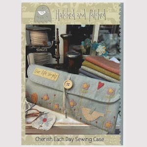 Puede incluir: Un estuche de costura azul claro con bordados florales y un diseño de pájaro, asegurado con un botón de madera. El texto "Live life simply" es visible. El estuche está etiquetado como "Cherish Each Day Sewing Case" y es de "Hatched and Patched."