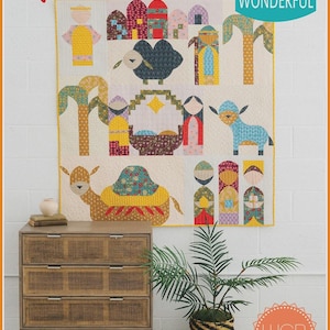 May include: A patchwork quilt depicting a nativity scene with figures, animals, and buildings. The word "Noel" is written in red script at the top. The quilt is displayed above a wooden chest of drawers with a plant in a woven basket.