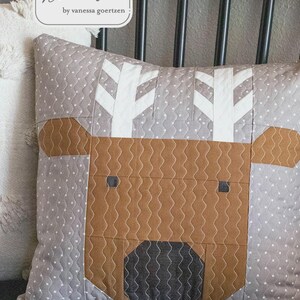 May include: A decorative pillow with a reindeer design. The pillow features a gray background with white polka dots and a stitched reindeer face in brown and black. The antlers are white. The text "Merry Reindeer" is visible.