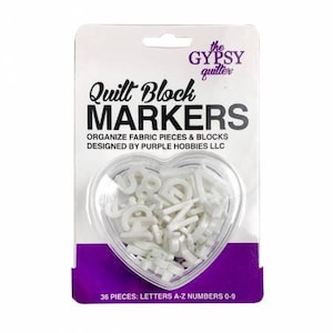 May include: White plastic letter and number markers in a heart-shaped container. The container is labeled "Quilt Block Markers" and "Organize Fabric Pieces & Blocks Designed by Purple Hobbies LLC". The container also says "36 Pieces: Letters A-Z Numbers 0-9".