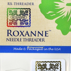 May include: A white card with blue and green accents featuring a needle threader. The card has the text "RX-THREADER" and "ROXANNE™ NEEDLE THREADER" in black text. The card also has the text "Made & Packaged in the USA" in blue text. The card has a small white square with a gold design and the text "ROXANNE" in gold text.