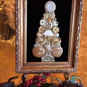 May include: A framed artwork featuring a Christmas tree made of buttons and other embellishments. The tree is decorated with gold, white, and brown buttons, and is set against a black velvet background. The frame is gold and ornate.