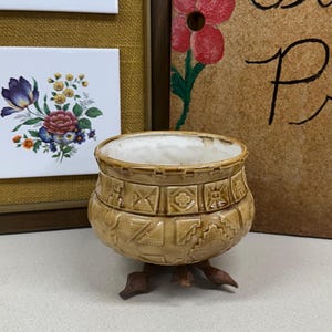 May include: A tan ceramic pot with a white interior, resting on a wooden base. The pot is decorated with geometric patterns. In the background, framed floral art and a sign that reads "Sophie Flower Press" are visible.