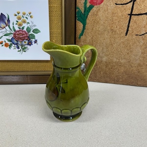 May include: A small, green ceramic pitcher with a handle and spout. The pitcher has a bulbous body with a decorative pattern and a dark green base. The pitcher is likely vintage and suitable for serving cream or other liquids.
