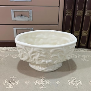 May include: A cream-colored ceramic bowl featuring a raised, ornate floral relief pattern around the exterior. The bowl has a wide, open top and sits on a pedestal base, showcasing a glossy finish and intricate sculpted details on its surface.
