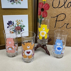 May include: Four vintage Care Bears drinking glasses with different bear designs. The glasses are clear with colorful illustrations of the bears. Two floral tile art pieces are in the background, along with a corkboard with the words "Flor Pre" written on it.
