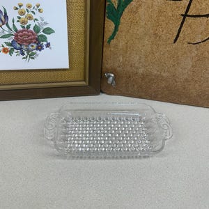 May include: A clear, rectangular glass butter dish with a textured interior and decorative handles. The dish is sitting on a light-colored surface. In the background are framed floral tiles and a wooden sign with a red flower.
