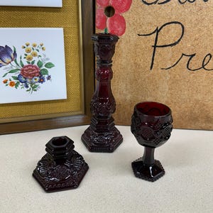 May include: Three pieces of ruby red glass tableware. Includes a small hexagonal candle holder, a tall candlestick, and a small goblet. The pieces have a detailed, embossed pattern.