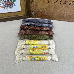 May include: A collection of raffia straw bundles in various colors, including cream, olive green, and brown. The image also shows a wooden sign with the words "Flower Press" and floral artwork, along with framed floral art.