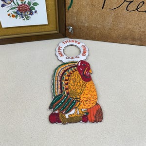 May include: A Thanksgiving-themed ornament featuring a colorful turkey with a red head, orange body, and green and orange tail feathers. The ornament has a white ring with the words "HAPPY THANKS GIVING".