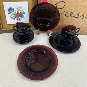 May include: A set of vintage dark purple glass dishes, including plates, saucers, and teacups. The dishes have a ridged design around the edges. The image also shows floral artwork and the words "Flower Press" on a wooden background.