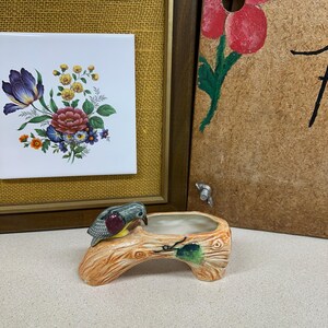 May include: A ceramic planter shaped like a tree branch, with a small bird perched on the edge. The planter is painted in shades of brown, green, and white. The bird has blue, red, and yellow accents. Decorative floral tiles are in the background.