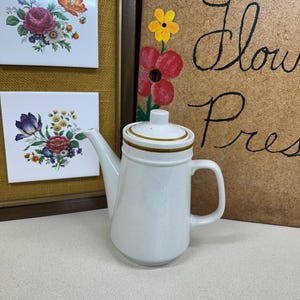 May include: A white ceramic teapot with a lid and handle. The teapot has a speckled finish and a brown band around the lid. Two framed floral tiles are in the background.