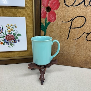 May include: A light blue ceramic mug with a handle sits on a small, dark wooden stand. The mug is cylindrical and has a smooth surface. In the background are framed floral tiles and a painted sign with flowers.