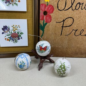 May include: Three white ceramic ornaments with floral designs. The ornament on the left has a blue ribbon and white flowers. The ornament in the center has a butterfly and green leaves. The ornament on the right has white flowers and green leaves.