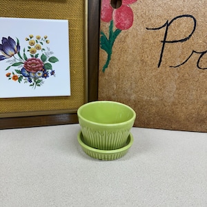 May include: A small, lime green ceramic planter pot with a matching saucer. The exterior of the pot features a vertical ribbed texture around the base, and the rim is smooth and rounded. The planter is a solid, bright color with a glossy finish.