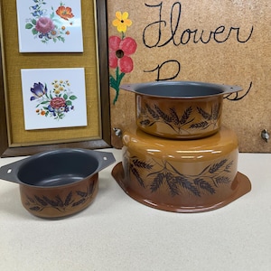 May include: Three-piece vintage cookware set in shades of brown with a wheat stalk design. The set includes a small pot, a medium pot, and a larger base. The pots have dark gray interiors and handles. The background includes floral artwork and the word "Flower".