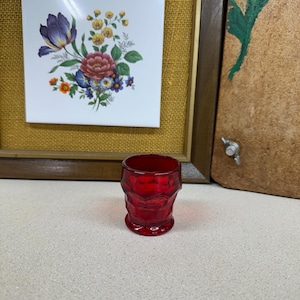 May include: A small, faceted, ruby-red glass. The glass has a textured design and a rounded base. In the background, a framed floral tile and a painted wooden panel are visible.