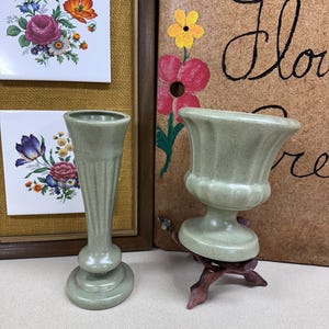 May include: Two green ceramic vases, one tall and slender with a flared top, the other a shorter, wider urn shape. Both vases are sitting on a table.