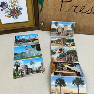 May include: A collection of vintage postcards featuring scenes from St. Augustine, Florida. The postcards display images of historic buildings, bridges, and palm trees. A framed floral print and a wooden press with handwritten text are also visible.