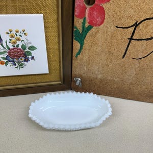 May include: A white, oval-shaped dish with a scalloped edge. The dish is made of opaque glass and sits on a light-colored surface. In the background, there is a framed floral tile and a wooden panel with a painted red flower.