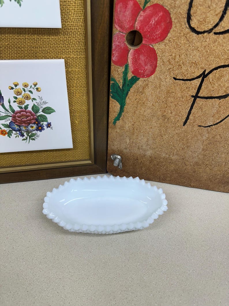 May include: A white, oval-shaped dish with a scalloped edge. The dish is made of opaque glass and sits on a light-colored surface. In the background, there is a framed floral tile and a wooden panel with a painted red flower.