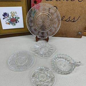 May include: Five clear glass plates with different patterns. The plates are arranged on a white surface. The largest plate has a floral design around the edge. The other plates have various geometric patterns.