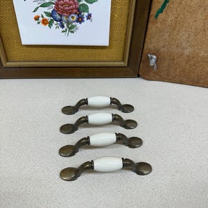 May include: Four antique-style cabinet pulls with white ceramic centers and bronze-toned metal hardware. The pulls are arranged in a row on a light-colored surface. The image also shows a framed floral print and a wooden panel.