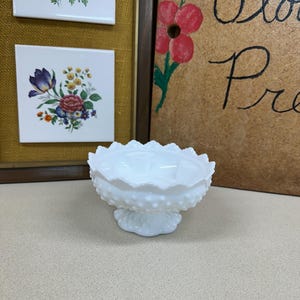 May include: A white milk glass pedestal bowl with a scalloped rim and a hobnail pattern. The bowl is on a pedestal base. Two framed floral tiles are in the background.