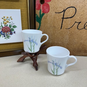 May include: Two white ceramic mugs with a blue iris flower design and a curved handle. One mug is on a small wooden stand. The background includes floral artwork and a sign with cursive writing.