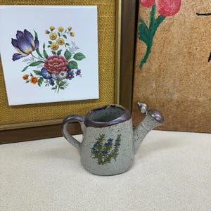 May include: A small, speckled ceramic watering can with a brown rim and handle. The body features a green and blue floral graphic. The piece has a short spout and a curved handle, showcasing a textured, off-white finish with dark brown speckles.