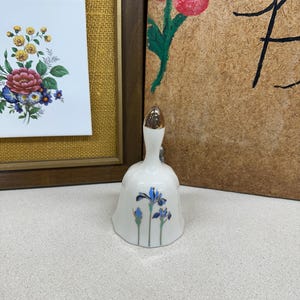 May include: A white ceramic bell with a gold-colored clapper and a floral design featuring blue irises. The bell is decorated with a gold rim and a gold-colored handle.