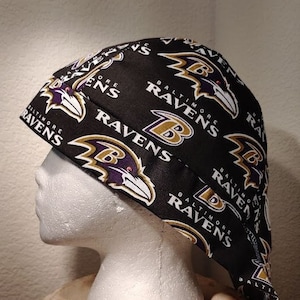 May include: Black and purple surgical scrub hat with a repeating pattern of the Baltimore Ravens logo. The logo features a raven with a purple and gold design. The text "Baltimore Ravens" is repeated throughout the hat.