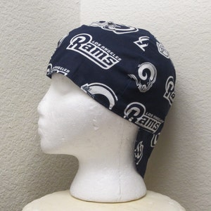 May include: A blue and white welding cap with the Los Angeles Rams logo repeated on the fabric. The cap has a flap that covers the neck.