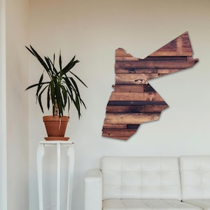 May include: A wooden wall art piece in the shape of Jordan. The wood is a dark brown color and has a rustic look. The art piece is hanging on a white wall in a room with a white couch and a plant in a terracotta pot.
