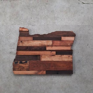 May include: A wooden wall art piece in the shape of the state of Oregon. The art is made of reclaimed wood planks in various shades of brown, creating a rustic and unique look.